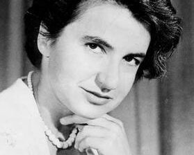 Black and white photo of Rosalind Franklin. She is wearing a pearl necklage and looking sideways at the camera with a slight smile. Her chin is on the fingers of her hand.